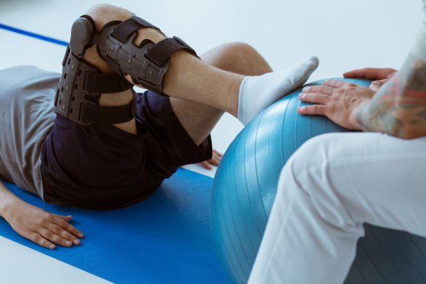 一个有腿部支撑的人在他们的瑜伽垫上躺在瑜伽垫上，右脚在瑜伽球上，别人持有稳定。