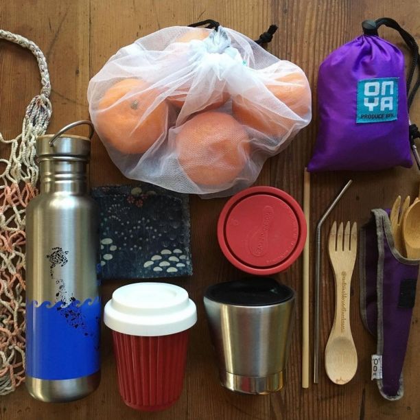 一个可重复使用的购物包放在木桌上，还有一个编织购物袋，金属水瓶，可重复使用的咖啡杯和切刀，以及可重复使用的农产品袋里的一袋橘子。