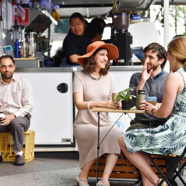 在一辆小咖啡车前，三个人坐在一张桌子前，用马克杯和可重复使用的咖啡杯喝咖啡，而背景里的一个男人用的是一次性塑料咖啡杯。